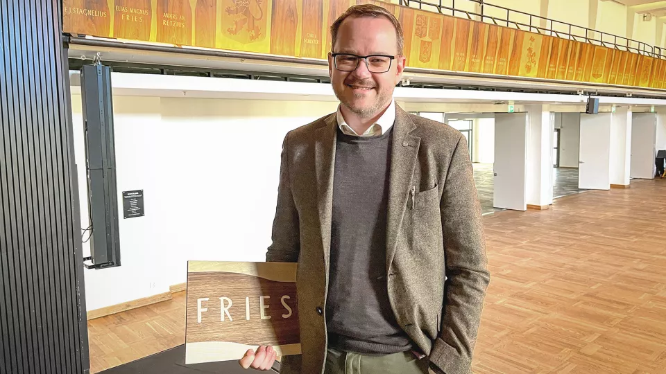 Foto av Johan Stenfeldt inne i Stora salen i AF-borgen i Lund. I handen håller han en bit trä gjort av intarsia.