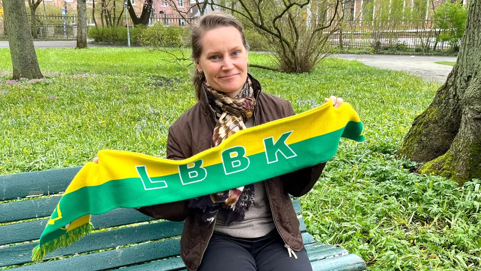 Porträttfoto av Lina Wedin som sitter på en bänk och håller upp en gröngul fotbollshalsduk.