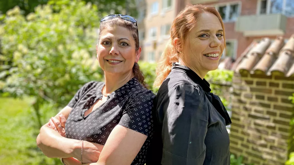 Foto av två kvinnor som står med ryggarna mot varandra och leende tittar in i kameran. 