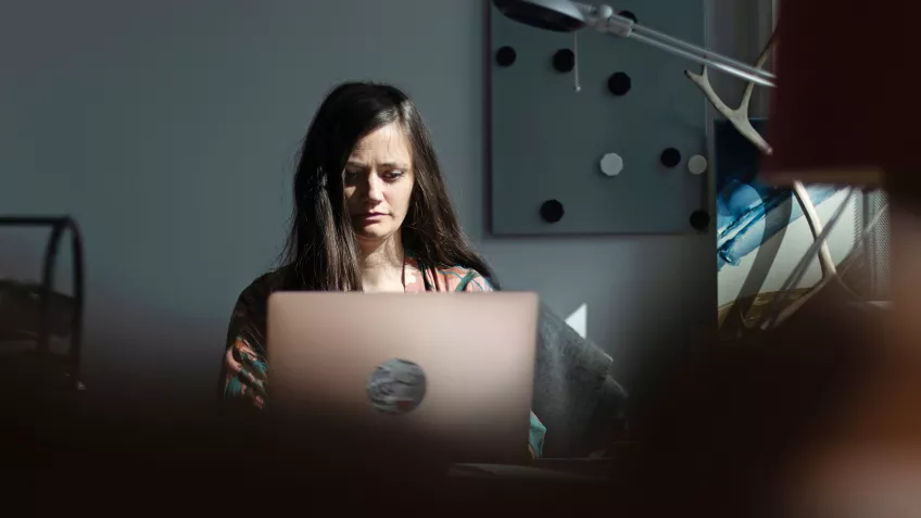 Foto av kvinna som sitter vid ett skrivbord och jobbar med sin uppslagna dator.